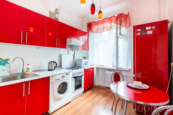 Кухня с красными шкафами, плитой, стиральной машиной и круглым красным столом с двумя стульями.