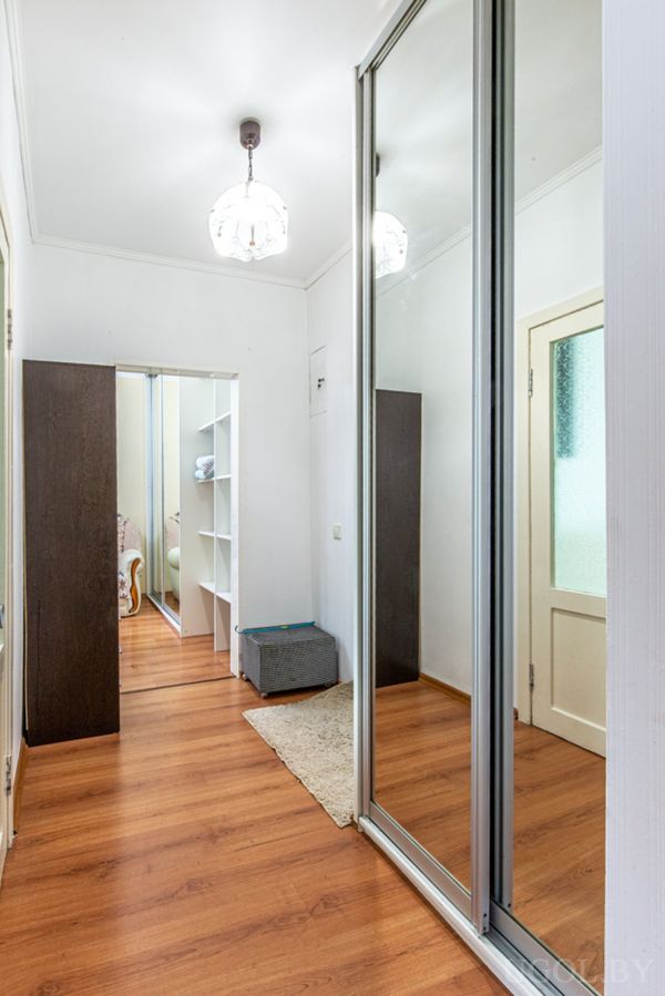 Светлый коридор с деревянным полом, зеркальным шкафом‑купе, белой дверью и тёмной полкой.