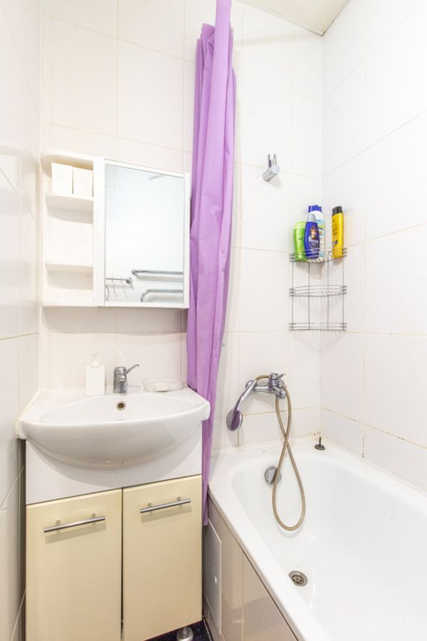 Небольшая ванная с белой тумбой, зеркальным шкафчиком, ванной с фиолетовой шторкой и полкой с шампунями.