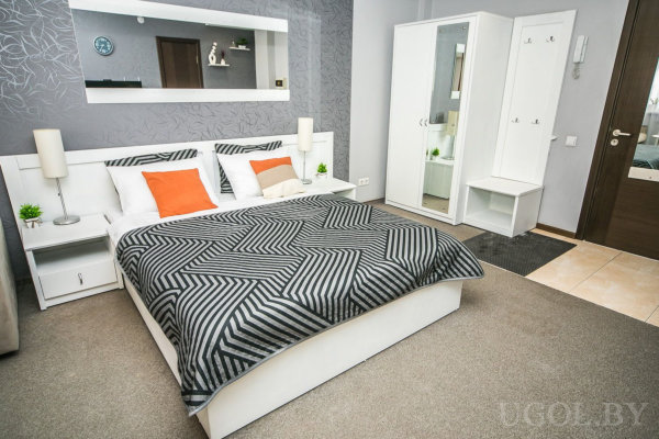 Белая кровать с чёрно-белым покрывалом и оранжевыми подушками, серый фон с узором и зеркалом, белый шкаф с зеркальными дверями и вешалкой, минималистичный интерьер.