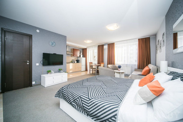 Кровать с геометрическим покрывалом и подушками, диван и столик, ТВ-зона с белой тумбой, кухня с деревянными шкафами и обеденным столом, большие окна с тюлем.