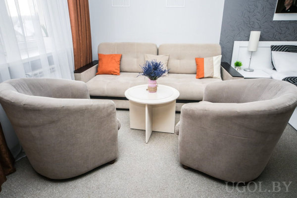 Бежевый диван с оранжевыми и белыми подушками, два кресла напротив, круглый столик с вазой и цветами, рядом видна кровать и прикроватная тумба.