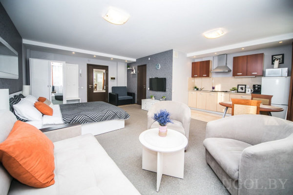 Два светлых дивана с оранжевыми подушками, белый столик с цветами, кровать с полосатым покрывалом, кухня с шкафами и техникой, обеденный стол с четырьмя стульями.
