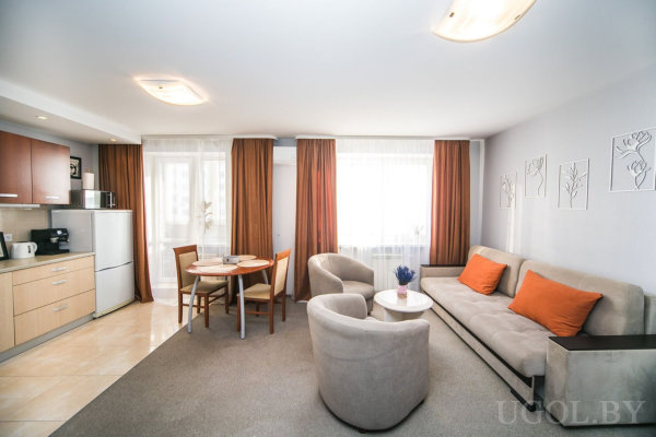 Кухня с деревянными шкафами и техникой, круглый стол с четырьмя стульями, диван с оранжевыми подушками, кресла и столик, большие окна с тюлем и шторами.