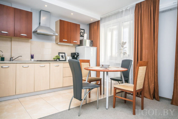 Нижние шкафы светлые, верхние тёмные, холодильник и микроволновка, стол с четырьмя стульями разного дизайна, большие окна с тюлем и шторами.