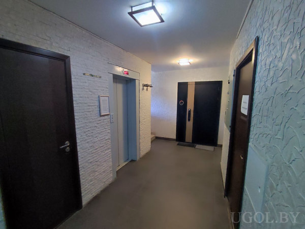 Лифт с красным индикатором, двери по сторонам, стены с белой кирпичной отделкой и штукатуркой, тёмный плиточный пол и современные светильники.