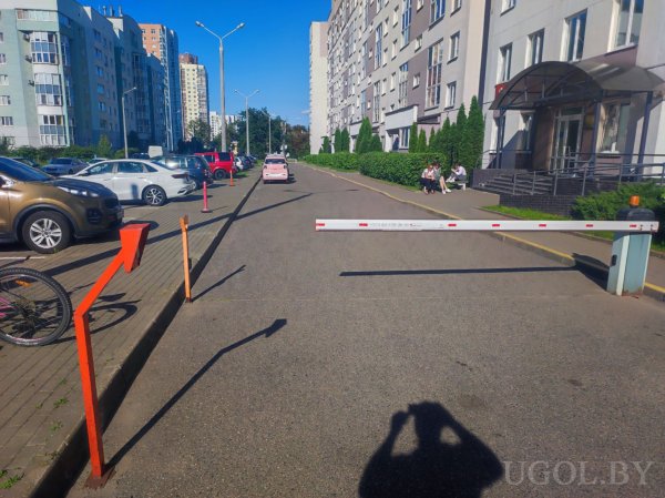 Белый шлагбаум с красными полосами, оранжевые стойки и указатель, припаркованные машины, люди на скамейке у подъезда, многоэтажные дома и зелень вдоль улицы.