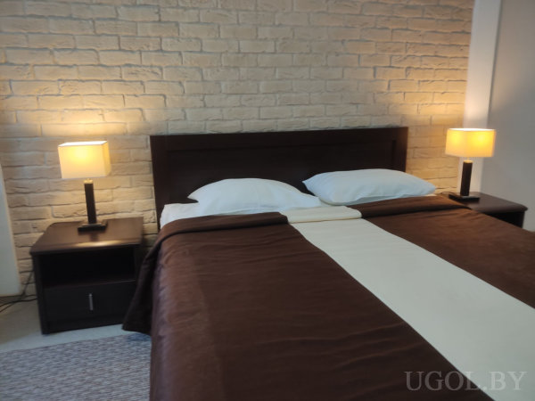 Кровать с коричневым покрывалом и светлым пледом, две тумбочки с лампами, светлая кирпичная стена и плетёный коврик у изножья.