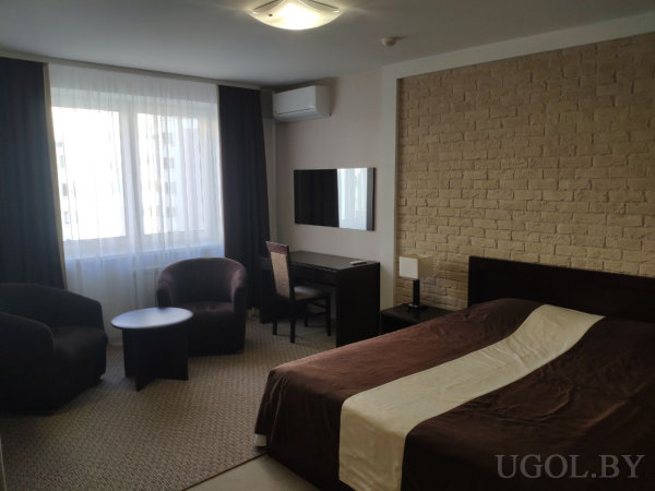 Кровать с коричневым текстилем, два тёмных кресла с круглым столиком, рабочий стол с зеркалом, большое окно с тюлем и плотными шторами.