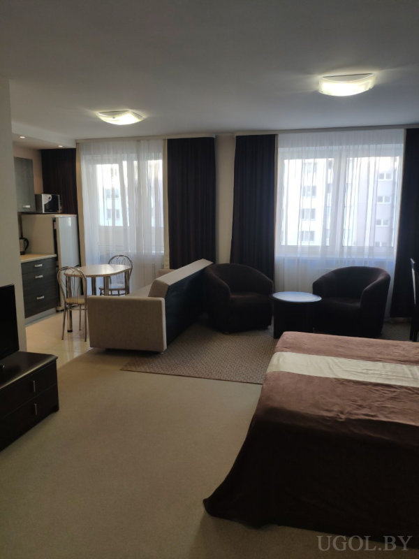 Кровать с коричневым покрывалом, два кресла с журнальным столиком, обеденная зона и компактная кухня с холодильником и микроволновкой.