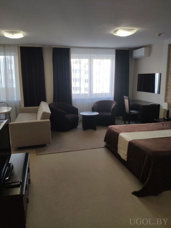 Кровать с коричневым текстилем, бежевый диван, два кресла, рабочий стол, два телевизора и большое окно с тюлем и шторами.