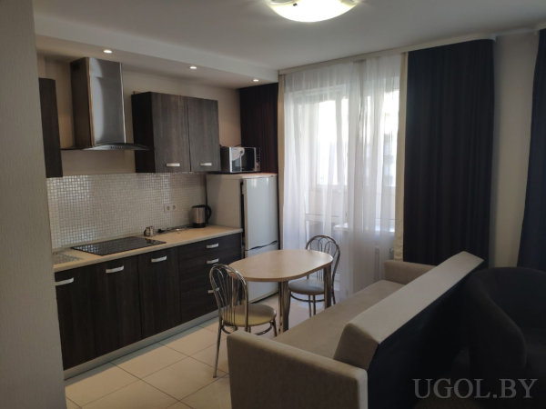 Кухня с тёмными шкафами, плитой и холодильником, рядом круглый стол с металлическими стульями и диван в бежево‑коричневой гамме.