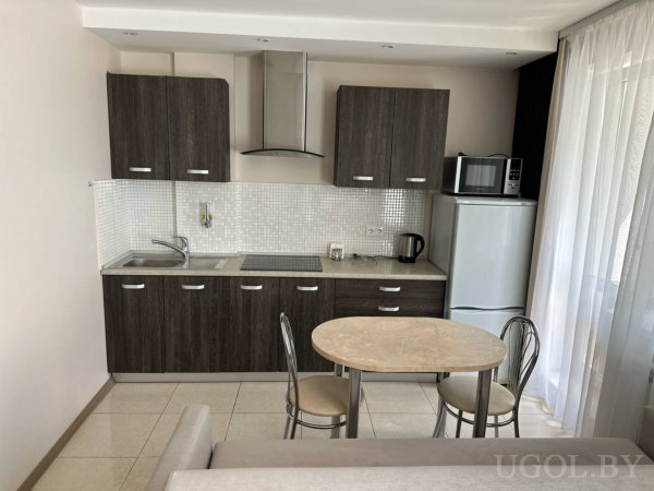 Тёмные шкафы, варочная панель, раковина, холодильник с микроволновкой, круглый стол с металлическими стульями и окно с тюлем.