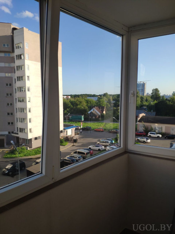 Панорамные окна лоджии, вид на многоэтажный дом, парковку, частный сектор и зелёные зоны, ясная погода.