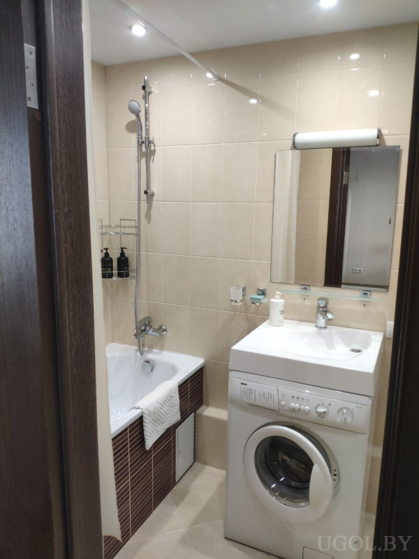 Белая ванна с душем, металлическая полка с чёрными бутылками, стиральная машина, раковина с зеркалом и бежевые плитки.