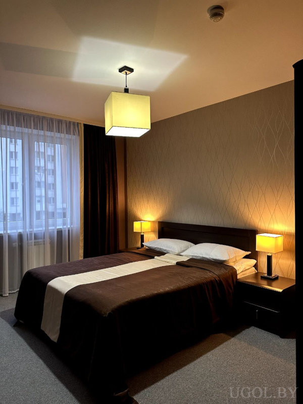 Двуспальная кровать с коричневым покрывалом и светлым раннером, две тумбочки с лампами, геометричные обои и окно с тюлем и тёмными шторами.