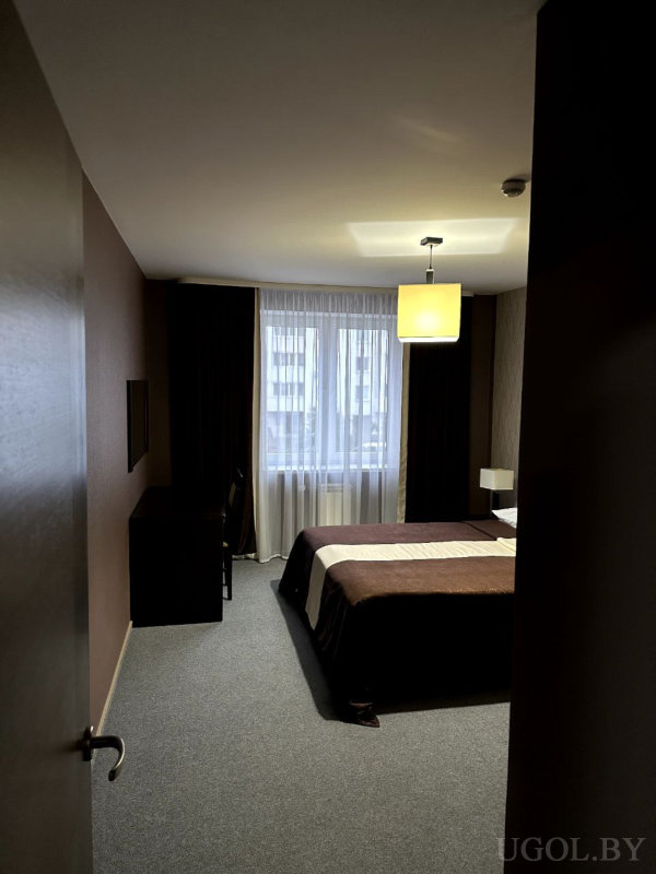 Кровать с коричневым покрывалом, рабочий стол с креслом, окно с тюлем и шторами, спокойная нейтральная палитра и мягкое освещение.