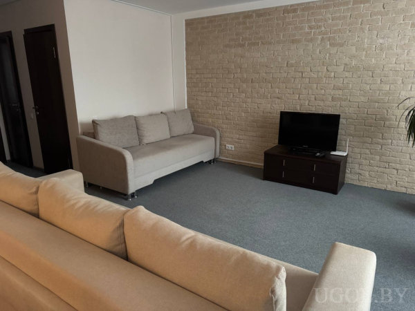 Два бежевых дивана, телевизор на тёмной тумбе у кирпичной стены, серый ковролин и две двери слева.