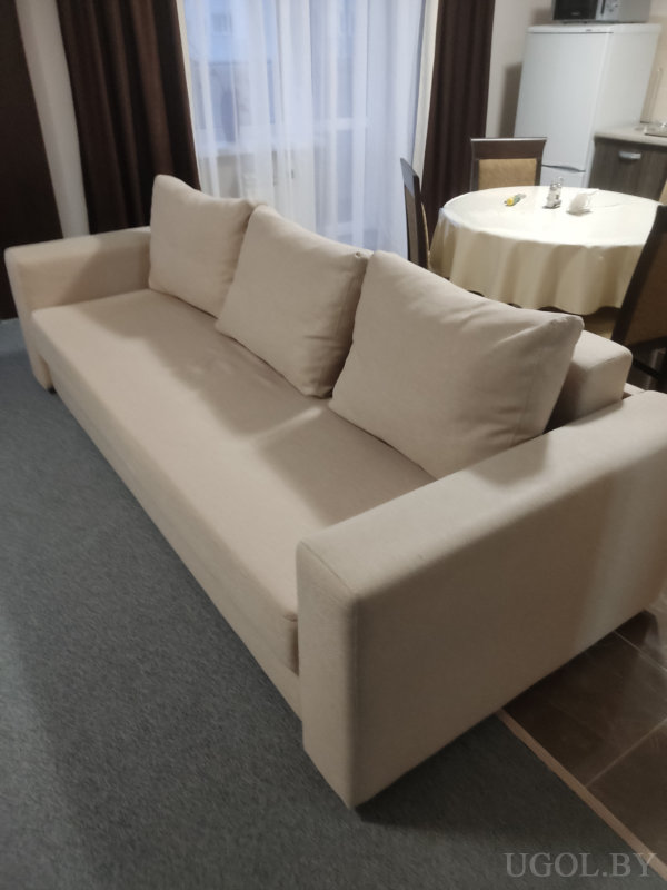 Бежевый диван с подушками, круглый стол с кремовой скатертью, окно с тюлем и шторами, рядом холодильник и часть кухни.