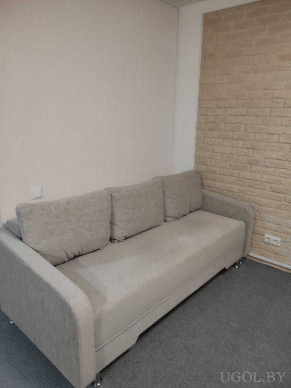 Серый диван с подушками, белая и кирпичная стены, серый ковролин и розетки по обе стороны дивана.