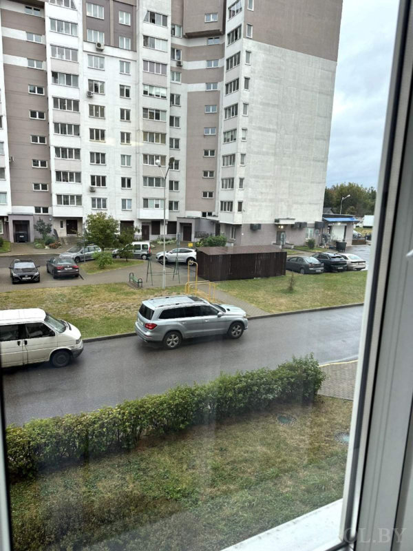 Многоэтажный дом с бело‑коричневым фасадом, зелёная зона, детская площадка, припаркованные машины и мокрый асфальт после дождя.