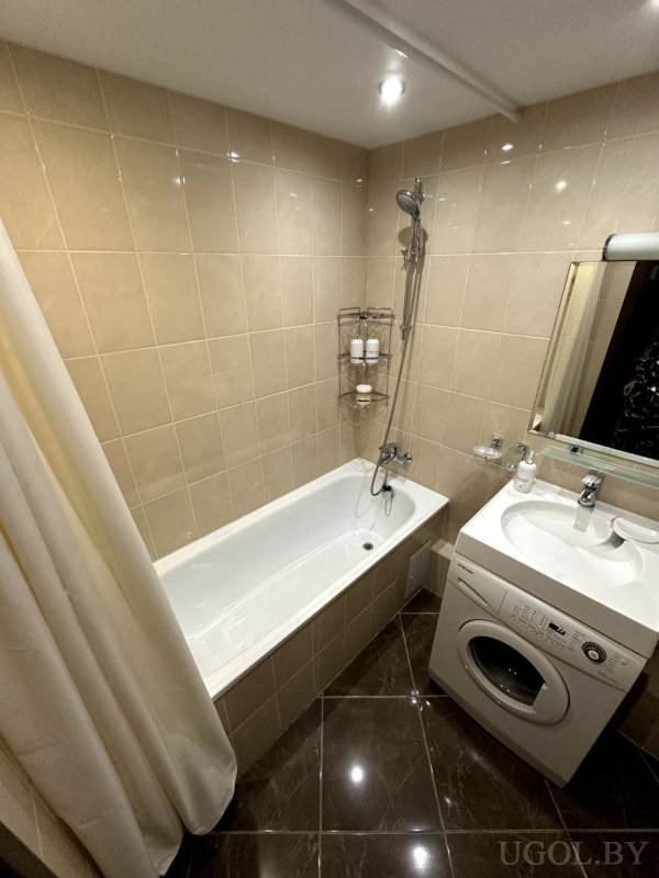 Белая ванна с душем, металлическая полка с косметикой, стиральная машина под столешницей, раковина с зеркалом и бежевые плитки.