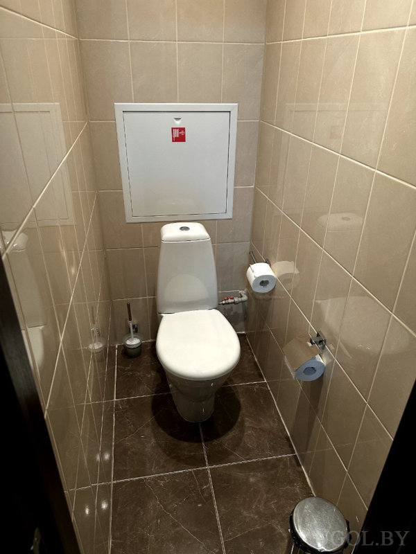 Белый унитаз у бежевой стены, три держателя туалетной бумаги, маленькая урна и доступный люк обслуживания.