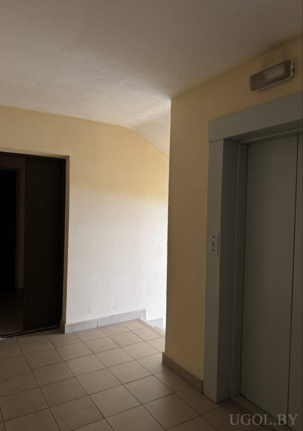 Лифт с серой рамкой, светлый кафельный пол, кремовые стены, настенный светильник и тёмный дверной проём слева.