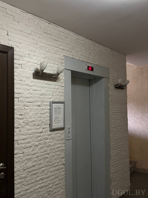 Лифт с цифровым индикатором этажа, белая кирпичная стена, два настенных светильника, кнопочная панель и дверь в коридор.