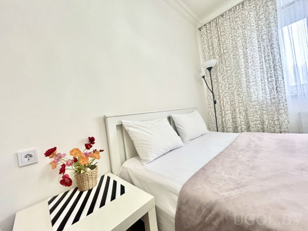 Белая кровать с розовым пледом, тумбочка со корзиной искусственных цветов, торшер и шторы с цветочным узором.