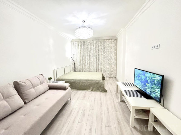 Кровать с зелёным покрывалом, бежевый диван, телевизор с природным сюжетом, люстра и напольная лампа на фоне светлых стен.