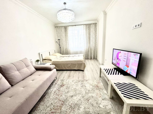 Бежевый диван с пуговичной стяжкой, кровать с бежевым покрывалом, стол с телевизором, комнатное растение и узорные шторы.