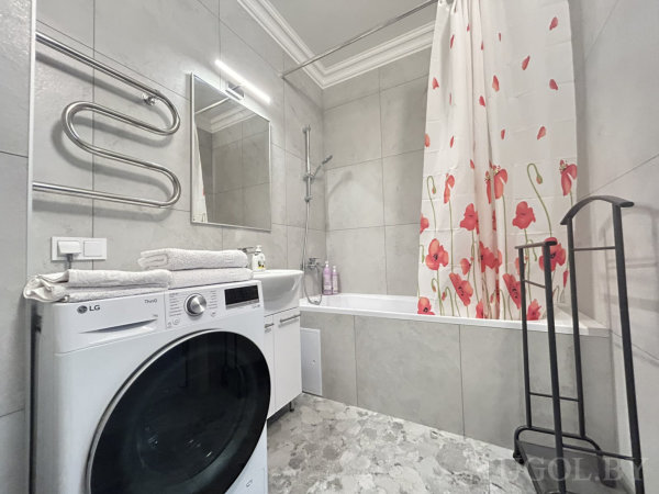 Стиральная машина под столешницей, зеркало с подсветкой, полотенцесушитель, ванна с занавеской с маками и серые плитки.