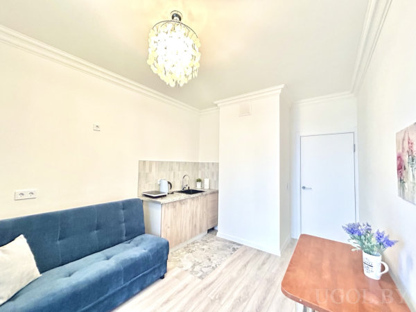 Синий диван, компактная кухонная зона с деревянным фасадом, стол с вазой и люстра, картина с цветами на белой стене.