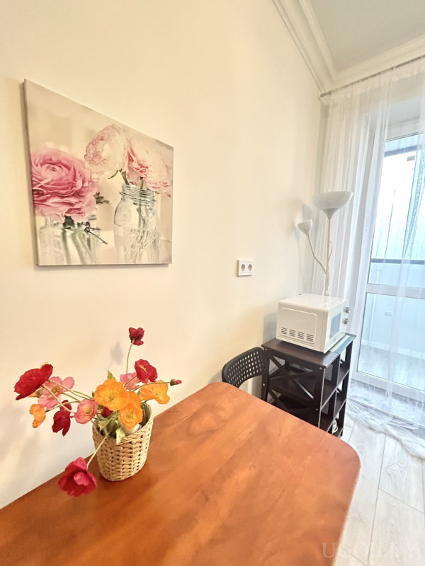 Деревянный стол с корзиной ярких цветов, картина с букетом, микроволновка на стеллаже, торшер и дверь с тюлем.