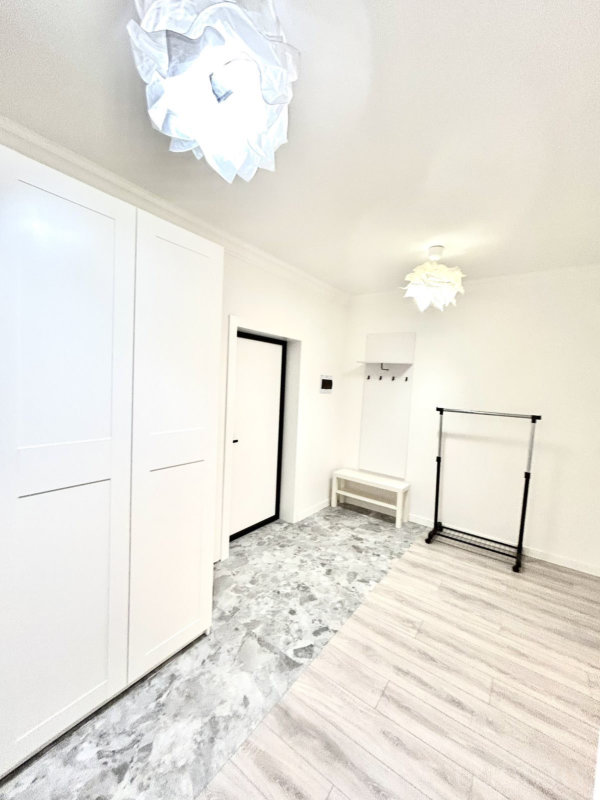 Белые стены и шкаф, скамья под настенной рейкой с крючками, чёрная стойка для одежды, серый кафель у входа и светлый ламинат.