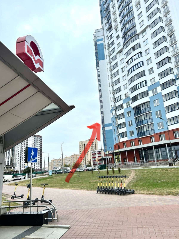 Высотный дом с красной стрелкой на фасаде, вход в метро, знак перехода и ряд электросамокатов на благоустроенной улице.