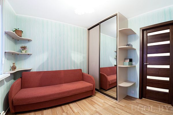 Небольшая комната с красным диваном, зеркальным шкафом‑купе и открытыми полками с декором.