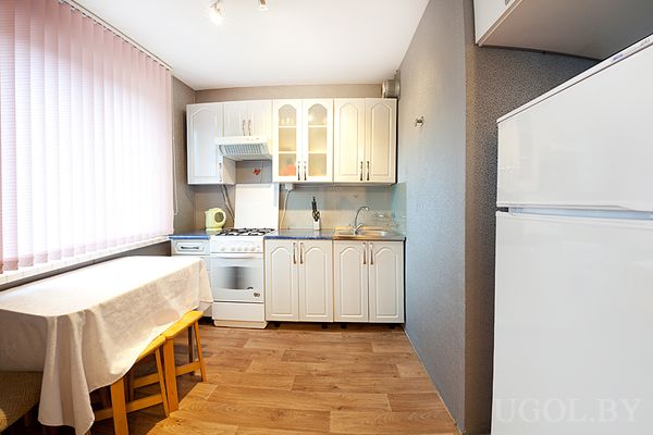 Компактная кухня с белыми шкафами, плитой, холодильником и столом у окна с розовыми жалюзи.