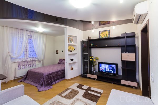 Комната с кроватью, шторой, полками, телевизором, тумбой, ковром и глянцевым потолком.