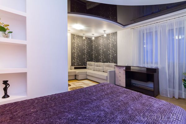 Интерьер с кроватью, диваном, столом, шторами, полками, тёмной стеной и точечным светом.