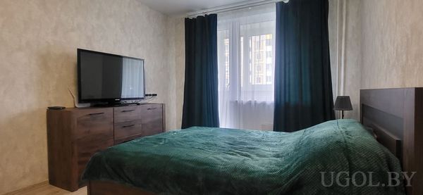 Комната с кроватью, деревянной тумбой, телевизором, торшером и шторами в тон покрывала.