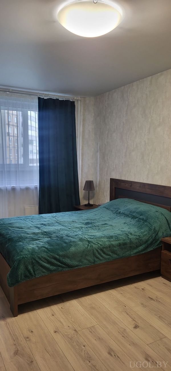 Интерьер с кроватью, зелёным покрывалом, деревянной тумбой, лампой и окнами с тюлем и плотными шторами.