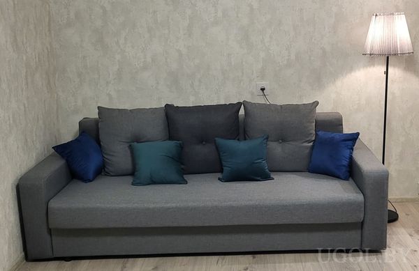 Комната с диваном, подушками в серо-синих тонах, торшером с плиссированным абажуром и светлой отделкой стен.