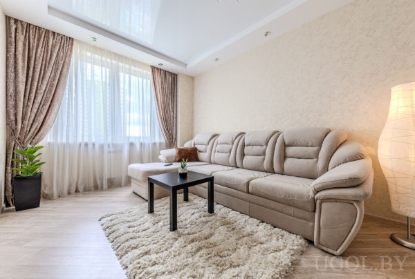 Светлая гостиная с большим бежевым диваном, пушистым ковром, чёрным столиком, высоким растением и окном с тюлем и коричневыми шторами.