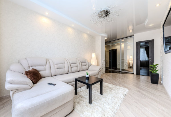 Комната с бежевым диваном, белым ковром, чёрным столиком, зеркальными дверями шкафа, тёмной дверью и телевизором на стене.