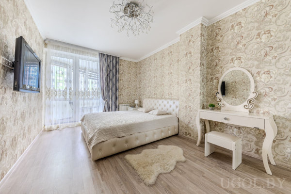 Спальня с кроватью с мягким изголовьем, бежевым покрывалом, белым туалетным столиком с круглым зеркалом, пушистым ковром и большими окнами с тюлем.