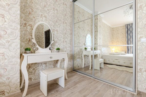 Белый туалетный столик с овальным зеркалом, декоративными растениями и зеркальным шкафом, отражающим кровать с мягким изголовьем.