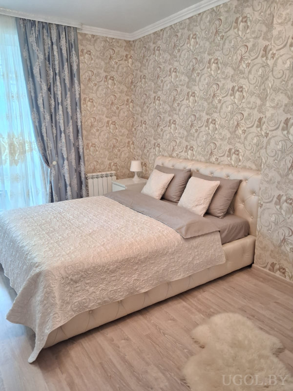 Кровать с кремовым изголовьем и бежевым текстилем, тумба с лампой, пушистый ковёр и окно с тюлем и синими портьерами.