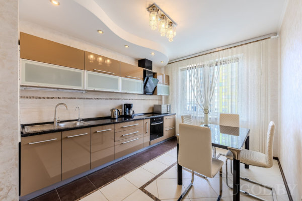 Современная кухня с коричневыми нижними шкафами, стеклянным столом с мягкими стульями, мраморным фартуком и потолком со встроенной подсветкой.